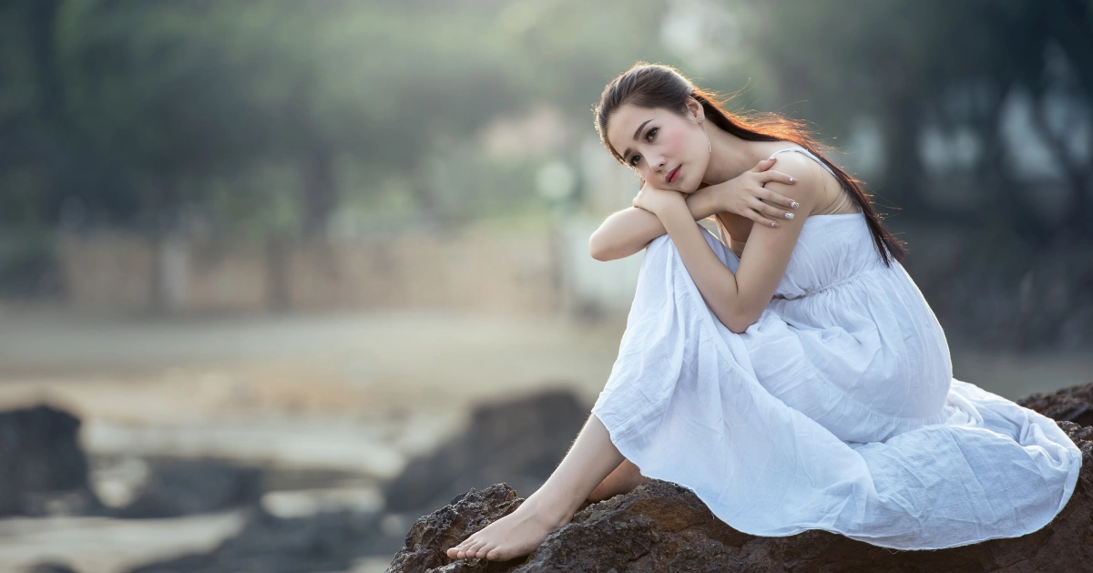 A woman in a white dress sitting alone at the beach.